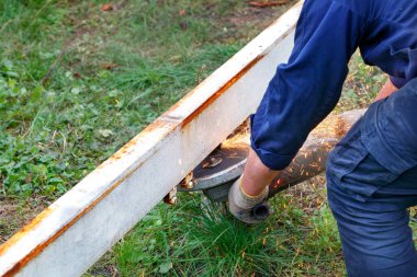 Mavi tulumlu bir işçi, çim biçme makinesiyle metal bir kirişi kesiyor, yeşil çimenlerin arka planına kıvılcımlar saçıyor. Boşluğu kopyala.