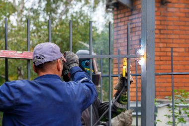 İnşaatçılardan oluşan bir ekip yeni evin etrafına metal bir çit yerleştirirken, biri ızgarayı zeminin altında tutuyor, diğeri de çiti kaynaklıyor..