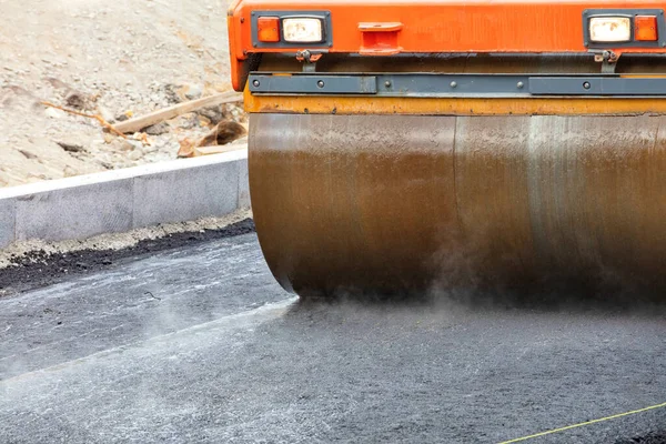 The metal cylinder of the large vibratory road roller compacts the fresh asphalt forcefully by being moistened with water against sticking on the road surface. Copy space, close-up.