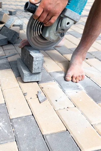 A worker with a grinder with a diamond cutting wheel trims paving slabs, standing barefoot and holding a stone cobblestone with one foot. Copy space, vertical shot.