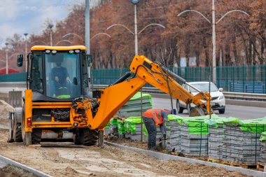 Çalışan bir ekip yol kazıcısını kullanarak kaldırım döşemeleri için yol kenarındaki döşemeleri boşaltıyor..