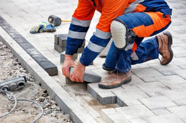 Mavi ve turuncu tulumlu, çömelmiş, çalışan bir tuğla ustası, elleri yeni bir yol üzerinde asfalt döşeyiyor. Yakın çekim, seçici odak, kopyalama alanı.