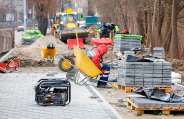 Yorgun bir müteahhit, akıllı telefonuna bakar ve bir bahar günü kaldırım döşemesi döşerken bir inşaat el arabasında bir çalışma alanının arka planında oturur ve arada dinlenir..