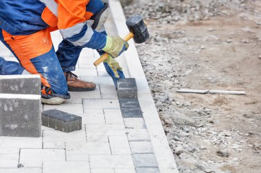 Mavi ve turuncu tulumlu bir işçi, yarı çömelmiş, kaldırım levhalarına plastik bir çekiçle vuruyor. Yakın çekim, seçici odak, kopyalama alanı.