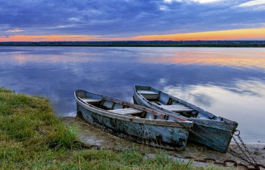 İki eski mavi ahşap tekne sakin bir nehrin kıyısına zincirlenir. Sabahın erken saatlerinde, yükselen güneş ışınları su yüzeyine ve bulutlu gökyüzüne yansıyor..