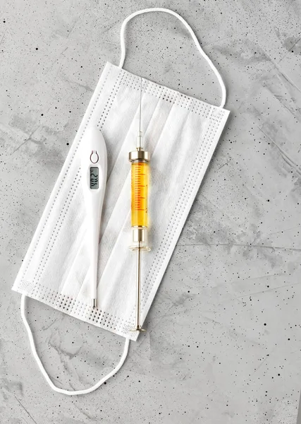 Medical protective mask, electronic thermometer and a syringe with a yellow vaccine on a gray concrete background. The concept of control and protection against viruses, flu and high body temperature. Vertical image, copy space, top view.