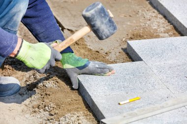 Mavi tulumlu bir işçi, yarı çömelmiş, granit bloklarda plastik çekiçle kapıyı çalıyor, yeni bir kaldırım yapıyor. Yakın çekim, seçici odak, kopyalama alanı.