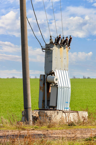 An old transformer station with bare wires stands near a concrete pole and a green agricultural field against a blue cloudy sky. Vertical image, copy space.