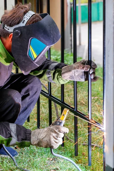 A welder in protective clothing, a helmet and leather gloves, using an electrode, welds a metal fence, installs a fence. Vertical image, selective focus, copy space.