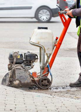 Yansıtıcı yelek giyen bir yol işçisi, şehir caddesinin önündeki eski asfaltı bulanık manzaralı bir benzin kesicisiyle kesiyor. Dikey resim, boşluğu kopyala.
