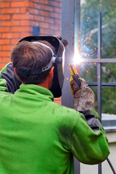 Kaynakçının iş yerinde parlak bir kaynak yayı koruyucu bir kask ve eldivenle hafif bulanık bir şekilde, çitin metal kısımlarını elektrot kullanarak kaynaklıyor. Seçici odak, dikey resim, boşluğu kopyala.