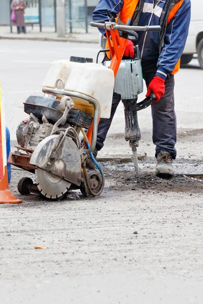 A road worker in reflective clothing on a fenced-in section of road repairs a roadway using an electric jackhammer and a gas cutter. Vertical image, copy space.