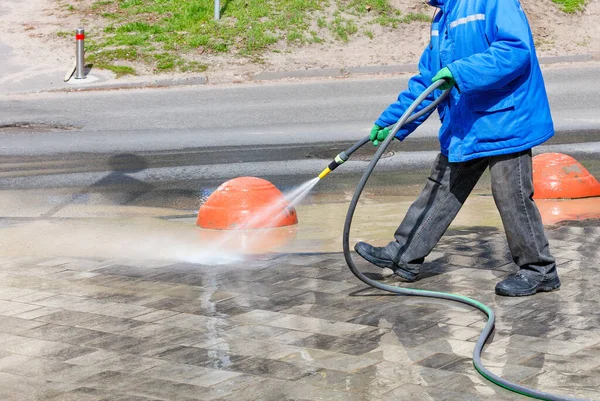 A janitor in a blue jacket pours water from a spray bottle on the sidewalk and flushes the dirt onto the side of the road on a clear, sunny day. Copy space.