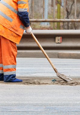 Turuncu yansıtıcı üniformalı bir hademe metal bir tamponun altındaki patikaların arasından kum topluyor. Dikey resim, boşluğu kopyala.