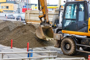 Bir yol kazıcı kovası kaldırımda çalışır. Kırmızı direkler, molozlar ve kumlar bir kamyonun arkasına konur. Şehir sokaklarının arka planına karşı..