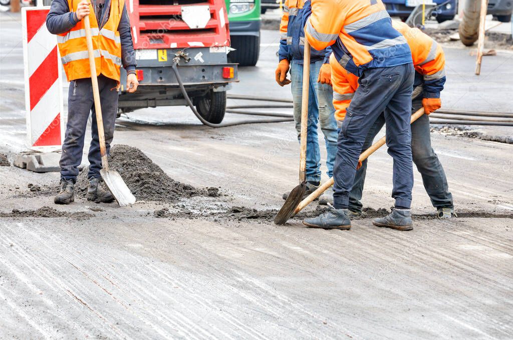 Road workers in orange overalls shovel debris from the old pavement on the roadway with shovels.