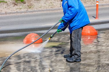 Mavi ceketli, su hortumlu bir hademe güneşli bir günde yol kenarındaki toprağı yıkıyor. Boşluğu kopyala.