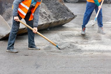 Parlak turuncu yansıtıcı yelekli yol işçileri eski yolu onarıyor ve yol döküntülerini bir kovaya kürüyorlar. Boşluğu kopyala.