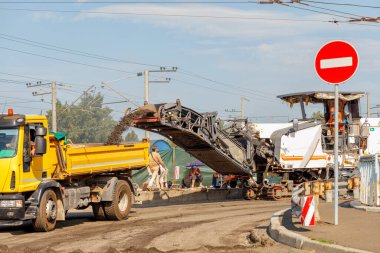 Sürüngen değirmen makinesi, tamir edilen yol kısmındaki asfalt tabakasını kaldırır ve yaz günü bir caddenin önündeki çöp kamyonuna yükler..