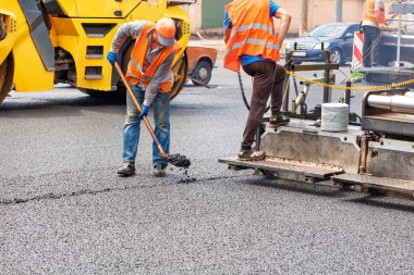 Yol işçilerinden oluşan bir ekip, asfalt kaldırım ve yol silindiri yardımıyla yolun bir bölümünü onarıp yola yeni bir sıcak asfalt tabaka seriyor. Boşluğu kopyala.