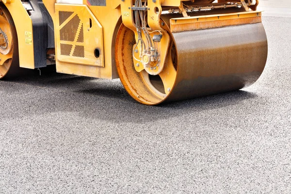 The metal cylinder of the yellow road vibrating roller compacts the fresh asphalt powerfully on the construction site. Copy space, selective focus, close-up.