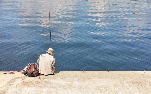 A fisherman in a light-colored panama hat and a vest sits on a concrete pier with a fishing rod near the water on a bright sunny day. Copy space.