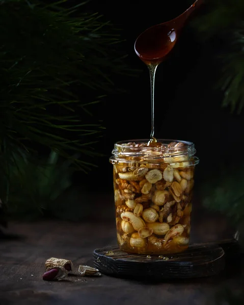 Nuts with honey in a glass transparent jar, a trickle of honey flows from a spoon, a dark wooden background, fir branches, close-up
