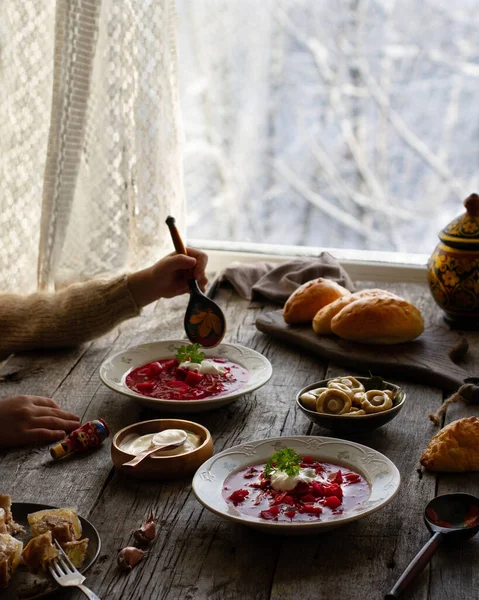 Traditional Russian lunch with borsch, mushrooms, jellied meat and pies, old wooden table, hands, Russian winter outside the window
