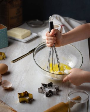 Bir çocuk şeffaf cam bardakta metal çırpıcıyla kurabiye hamuru yapar. Çocukların elleri yumurtayı şekerle çırpar..