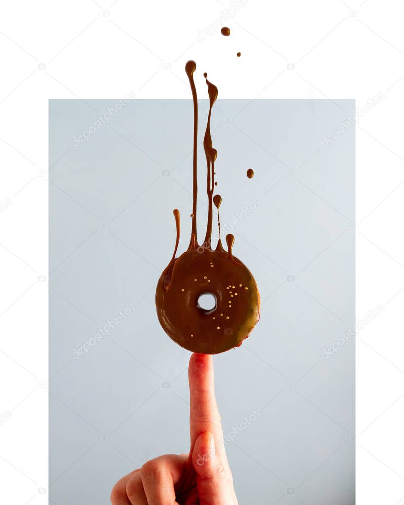 A chocolate donut drizzled with icing balances on the finger, the icing drips and drips up. Blue background.