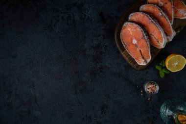 Koyu dokulu arka plan ve kenarları açık: tahtanın üzerinde somon bifteği, tuz, biber, yağ ve limon..