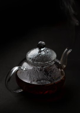 Siyah arkaplanda püskürtülmüş buharlı siyah çay ile karıştırılmış şeffaf cam çaydanlık