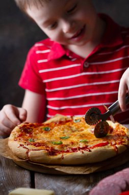 Bir çocuk pizza bıçağıyla yuvarlak bir pizzayı keser, gülümser, sevinir.