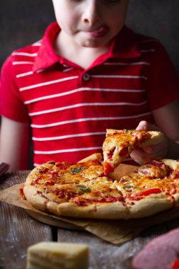 Bir çocuk domates ve peynirli bir dilim pizzayı deniyor, eliyle alıyor, dudaklarını yalıyor, yakın plan.