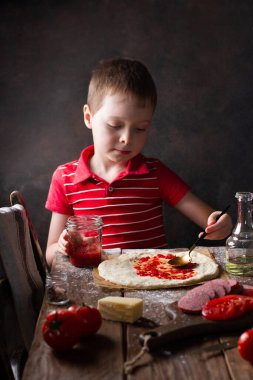 Çocuk, içinde malzemeler olan ahşap bir masada pizza hamuruna kaşık ile domates sosu sürüyor.