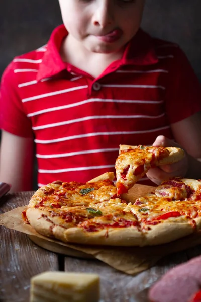 Bir çocuk domates ve peynirli bir dilim pizzayı deniyor, eliyle alıyor, dudaklarını yalıyor, yakın plan.