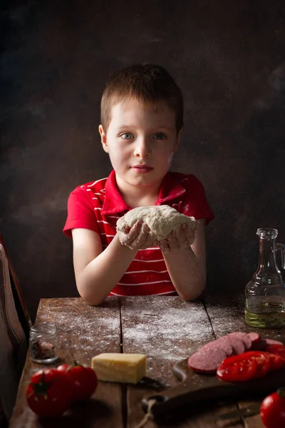 Bir çocuk hamuru iki eliyle mutfakta tutuyor. Ahşap bir masa ve malzemelerle, un üzerinden uçuyor.