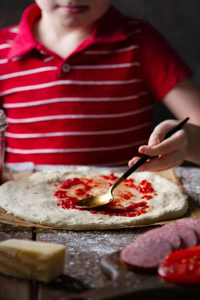Çocuğun eli, pizza tabağına domates sosu sürer. Parşömen üzerinde, tahta bir masada, yakın plan bir kaşıkla.