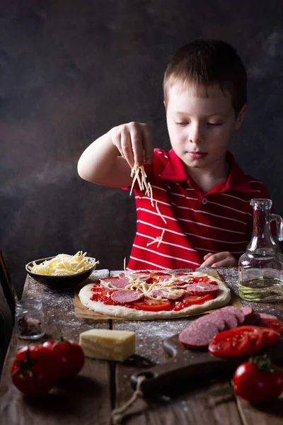 Çocuk mutfakta, ahşap kahverengi bir masada pizza hazırlıyor, rendelenmiş peynir döküyor.
