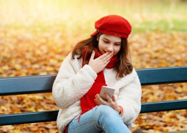 Kırmızı şapkalı ve atkılı kız telefona bakıyor ve merak ediyor ve sonbahar parkında bankta oturuyor.
