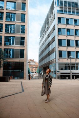 Kırmızılı kısa saçlı, gri elbiseli bir kadın tatilinde şehirde yürüyor.