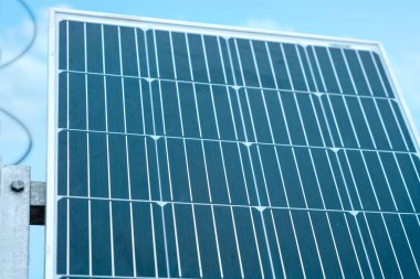 A close-up photo of a solar panel against a blue sky. The panel is made up of many small squares, and the sun is shining brightly.