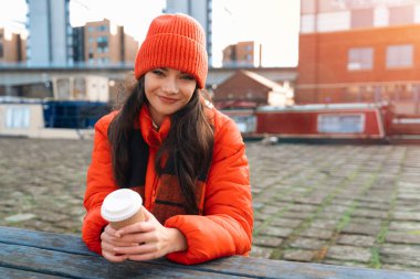 Genç bir kadın elinde bir fincan kahve, elinde kırmızı bir bere ve ceket, soğuk bir günde rıhtımda otururken gülümsüyor..