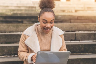 Kocaman gülümseyen bir kadın dışarıdaki taş basamaklara dizüstü bilgisayar kullanarak oturur. O bej bir ceket ve kazak giyiyor..