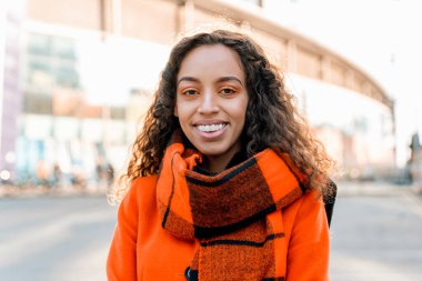 Turuncu bir ceket ve ekose bir eşarp giyen genç bir kadın dışarıda dururken gülümsüyor..