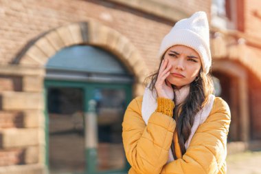 Beyaz bereli ve sarı ceketli genç bir kadın kışın dışarıda duruyor. Düşünceli bir ifadeyle aşağıya bakıyor ve eli yanağında dinleniyor..