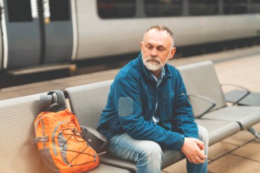 Tren istasyonundaki bankta oturan bir adam yanında turuncu bir sırt çantasıyla tren bekliyor..