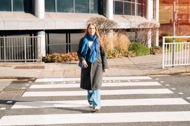 Uzun gri ceketli ve mavi eşarplı bir kadın modern bir binanın önündeki yaya geçidinden geçiyor..