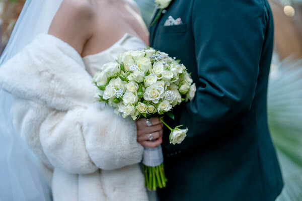 Невеста в белом меховом пиджаке держит красивый букет белых роз с зеленью, стоя рядом со своим женихом.