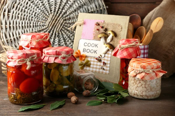 Jars with pickled vegetables and beans, spices, book of recipes and kitchen utensils on wooden background — Stock Photo, Image